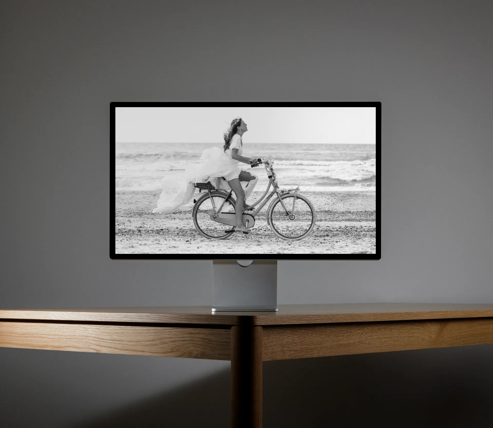 En computerskærm på et træbord viser et sort-hvidt billede af en kvinde i brudekjole, der cykler langs en sandstrand ved havet.