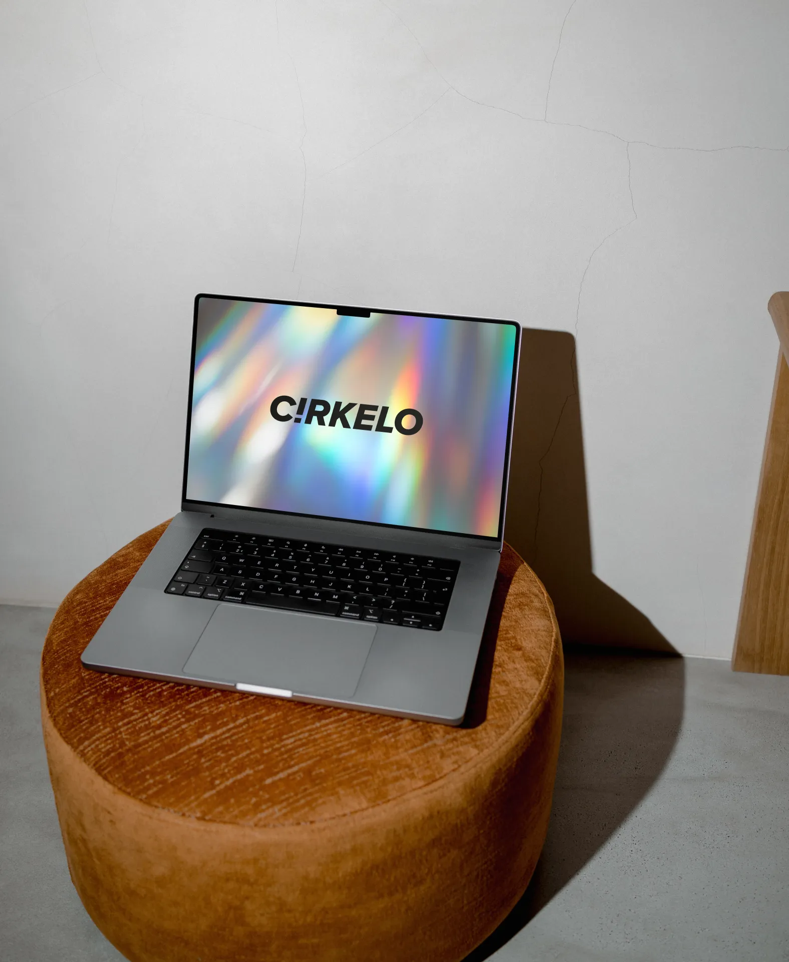 En sølv bærbar computer står på en rund puf i orange velour foran en hvid væg og viser C!RKELO-logoet på en farverig, iriserende baggrund.