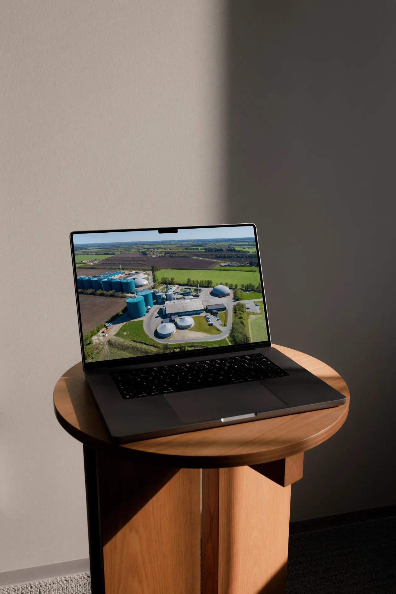 En mørk bærbar computer står på et rundt træbord og viser et luftfoto af Ribe BioGas med blå lagertanke omgivet af grønne marker.